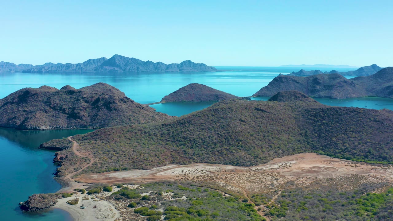 At 0:03, the aerial view shows brown-green hills sloping to a curved Juncalito shore, a blue inlet at left, distant mountain islands across the water, and no visible people.