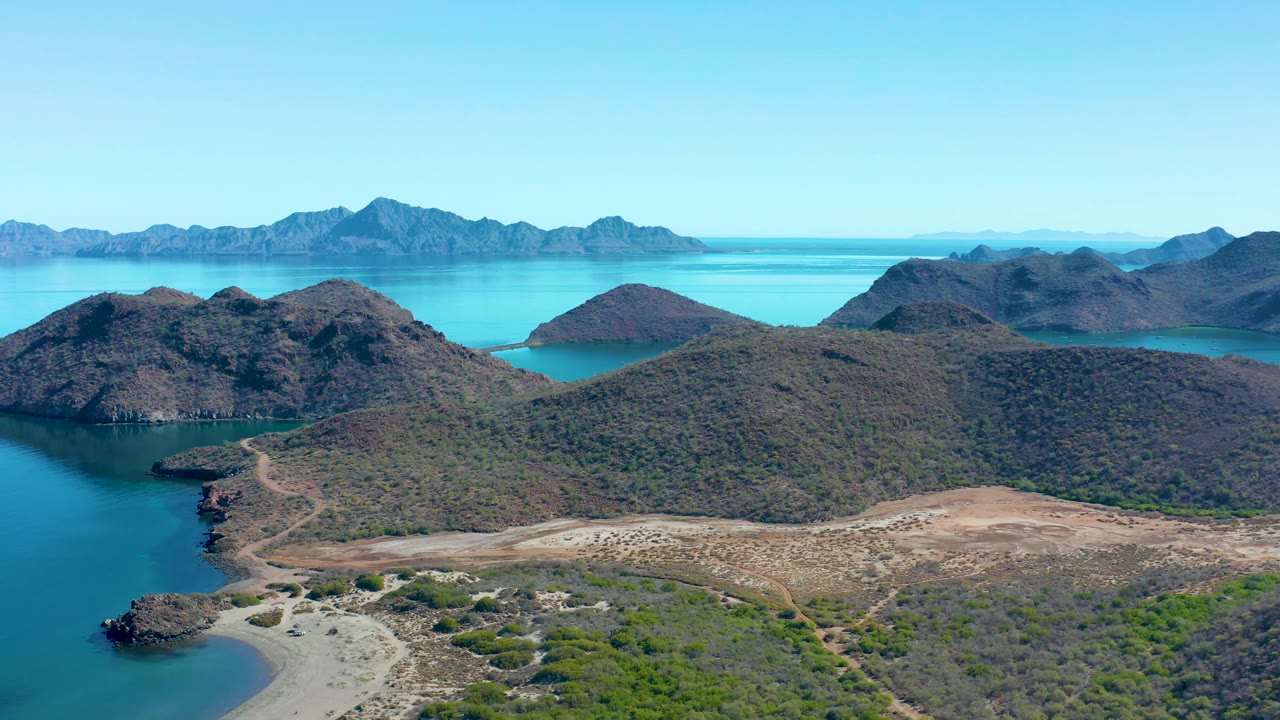 Wide drone view of turquoise water, dry shoreline paths, scrub hills, and mountain ridges.
