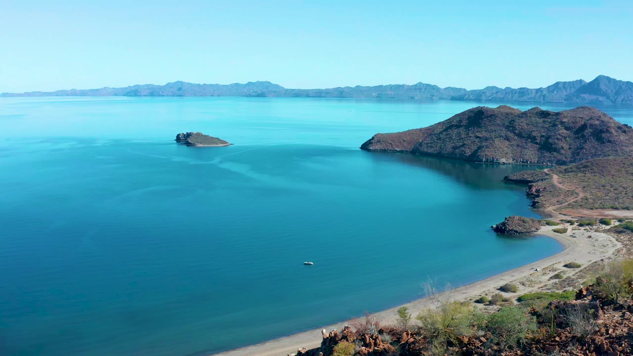 Wide Juncalito aerial frame showing calm blue-green water, a small island left of the rocky headland, sandy shoreline at lower right, distant mountains, and no visible people.