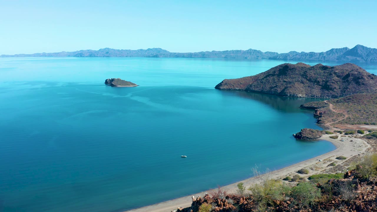 Aerial frame over the Juncalito bay with the rocky hill mass on the right, a small island offshore, pale beach and scrub at the bottom edge, distant mountains, and no visible people.