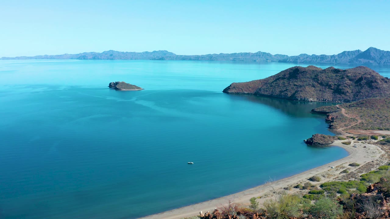 Juncalito aerial view with calm water across most of the frame, a curving beach and greener shoreline vegetation more visible at lower right, rocky hills, distant mountains, and no visible people.