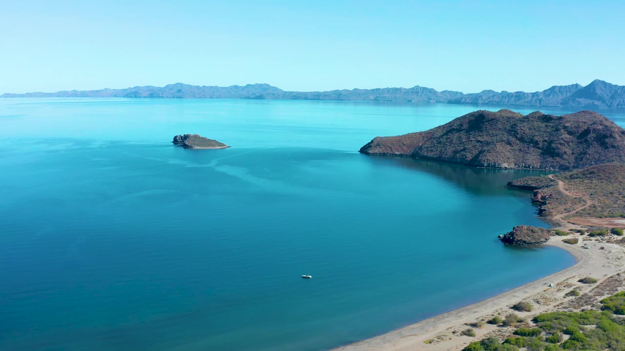 At 0:01, the Juncalito aerial view keeps the small boat low in frame while a rocky island, curved beach, blue water, and mountain horizon remain visible, with no visible people.