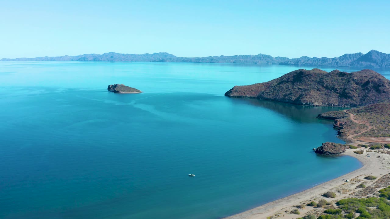 At 0:02, the boat sits farther into the open blue water of Juncalito bay, with the sandy beach and rocky shoreline on the right and layered mountains across the horizon, with no visible people.