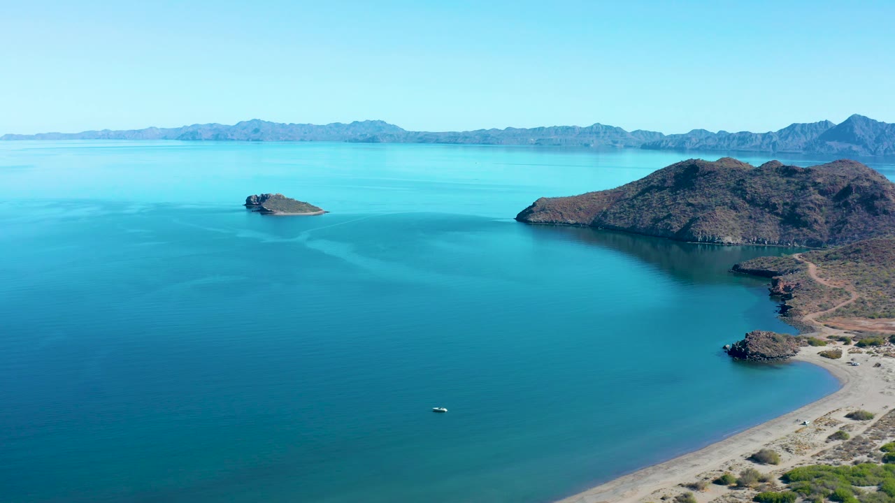 At 0:03, the aerial view shows the small boat low in the blue bay, a pale sand shoreline at right, rocky islands across the middle distance, and mountains beyond, with no visible people.