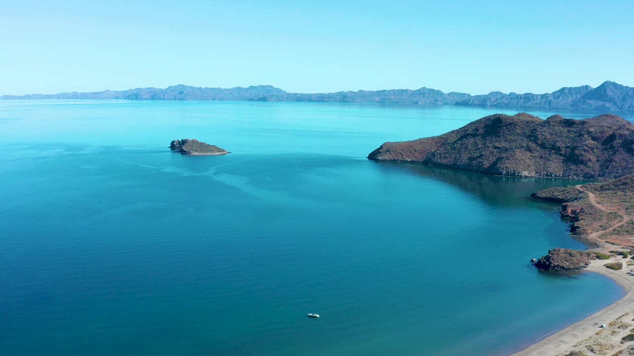 Aerial frame over Juncalito, Baja California Sur, showing turquoise bay water, a small rocky island left of center, a larger island to the right, distant mountains, and no visible people.