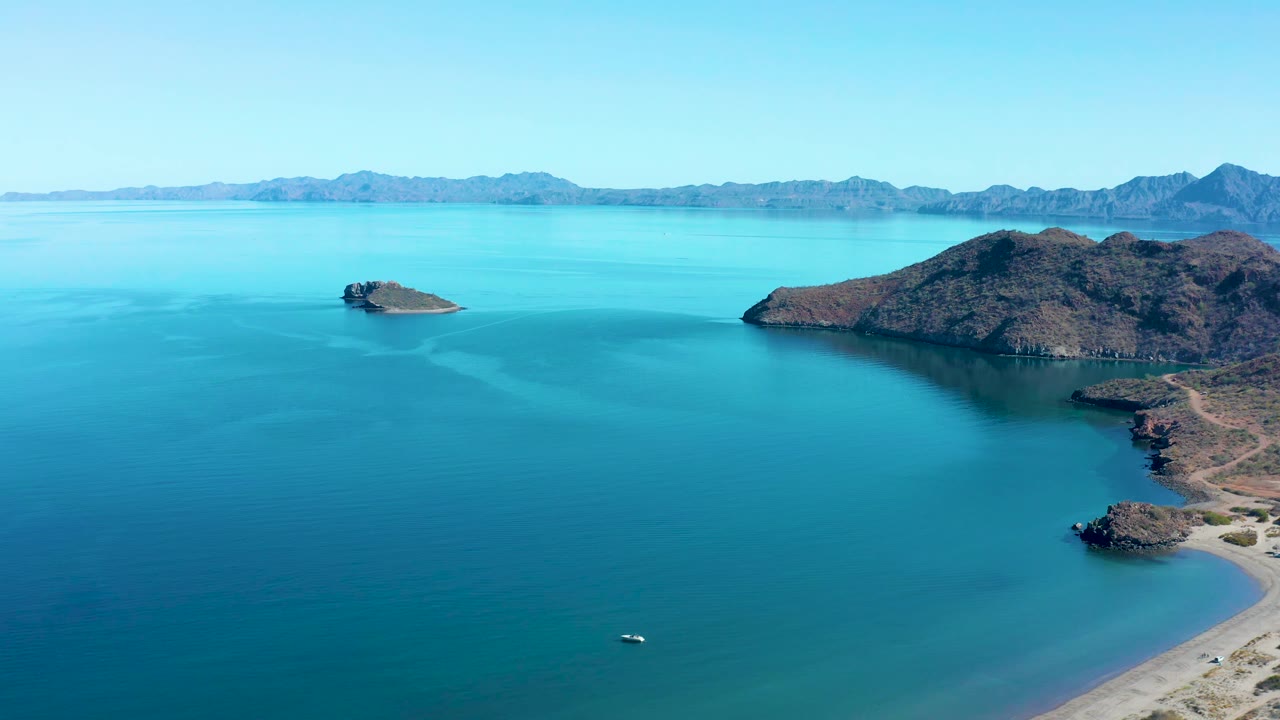 Second aerial frame over Juncalito shows the small rocky island slightly left of center, the larger island across the right midground, smooth blue water, a small boat low in frame, and no visible people.