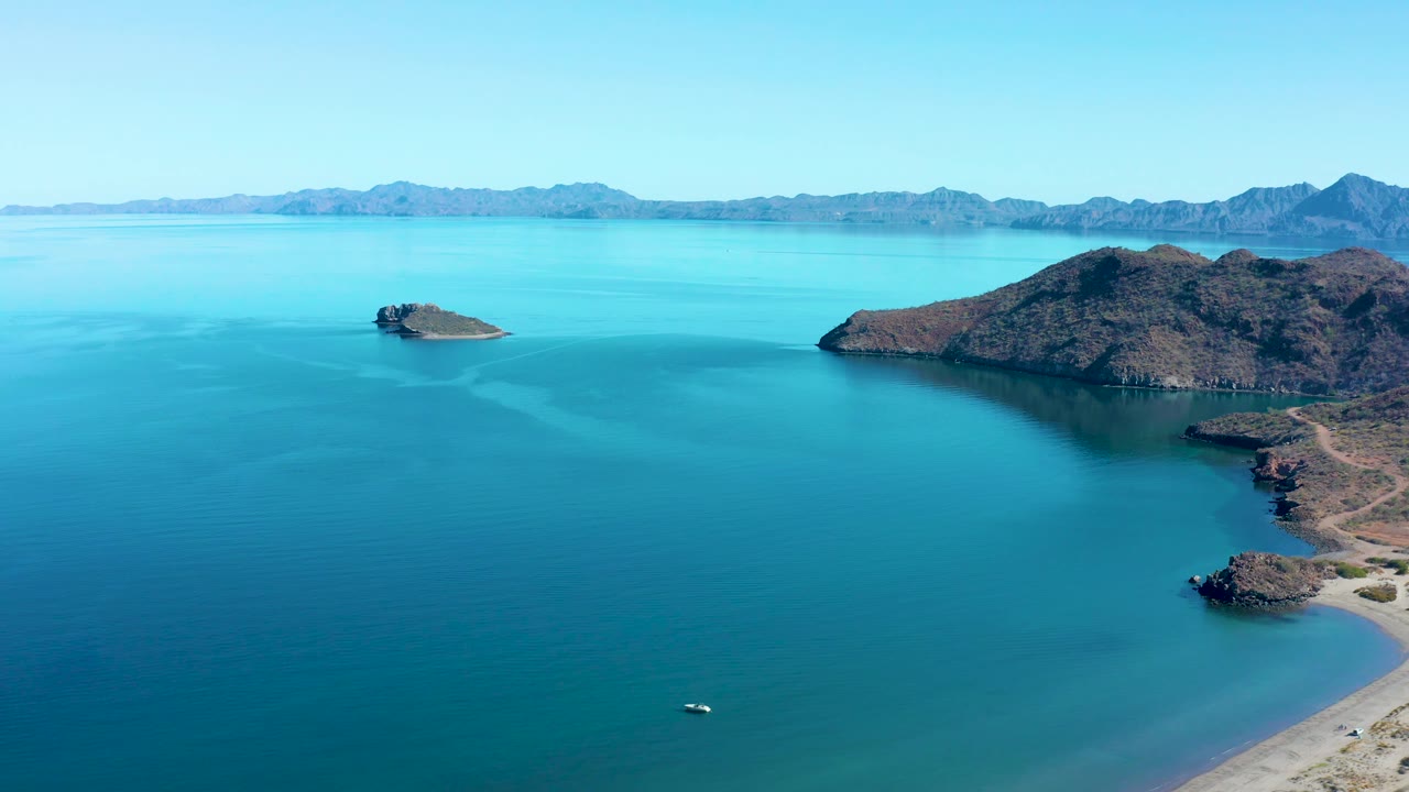 Aerial view in Juncalito with calm turquoise water in the foreground, a small rocky island near the left midground, a larger rugged island to the right, low mountains on the horizon, and no visible people.