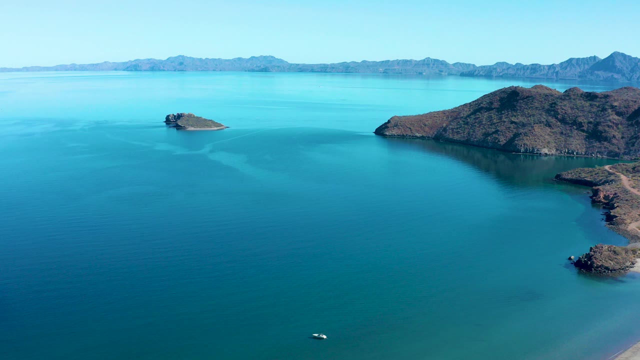 Aerial view of blue coastal water, rocky islands, distant mountains, and a small boat near the lower edge.