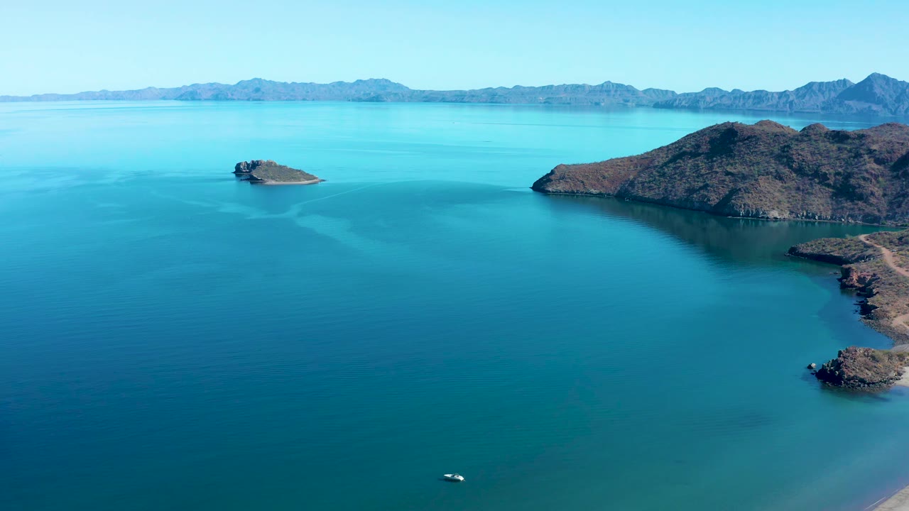 At 0:01, the Juncalito aerial angle holds on turquoise water, a small boat low in frame, a rounded rocky island left of center, larger rocky land to the right, distant mountains, and no visible people.