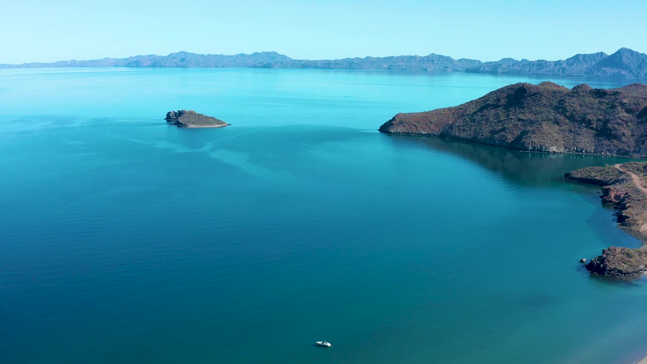 At 0:02, the Juncalito view emphasizes open blue water, rocky landforms to the right, a small island left of center, a mountain horizon, clear sky, and no visible people.
