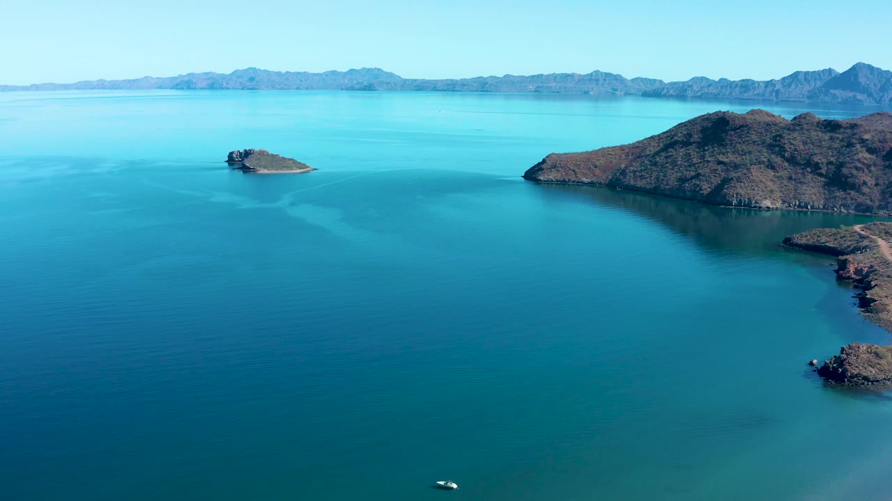 At 0:03, a small white boat sits near the bottom edge of the Juncalito aerial frame, with calm blue water, rocky islands to the right, distant mountains, clear sky, and no visible people.