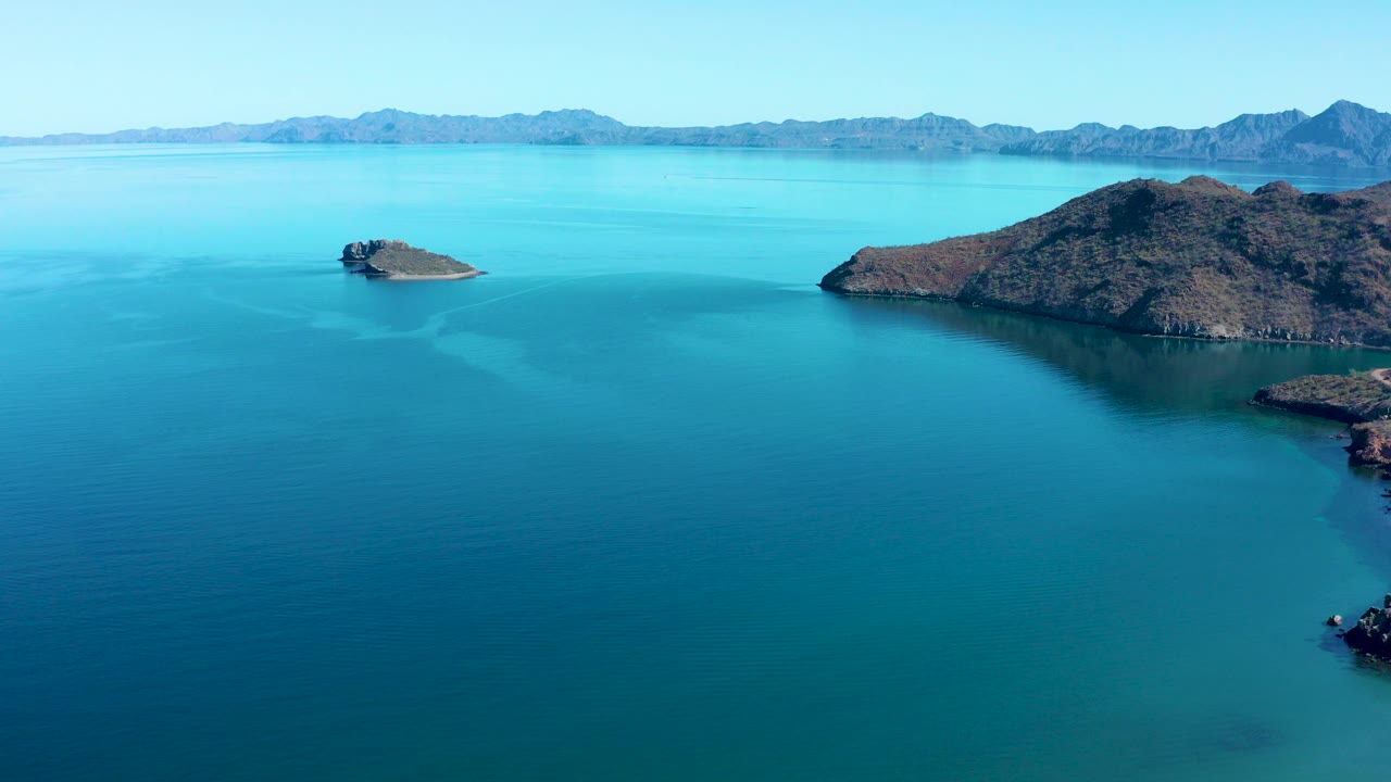 Aerial view over Juncalito, Baja California Sur, with turquoise water, a small rocky island left of center, a larger rocky landform on the right, distant mountains, and no visible people.