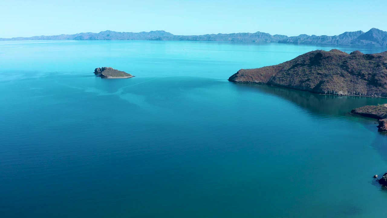 Aerial frame over Juncalito water with the small island left of center, the larger rocky mass at right, faint tonal bands in the water, distant mountains, and no visible people.
