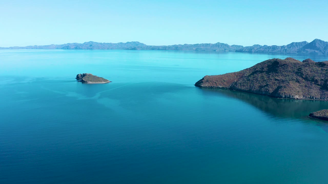 Opening frame over Juncalito, Baja California Sur, Mexico, showing turquoise sea, a small rocky island left of center, a larger island at right, distant mountain ridges, clear sky, and no visible people.