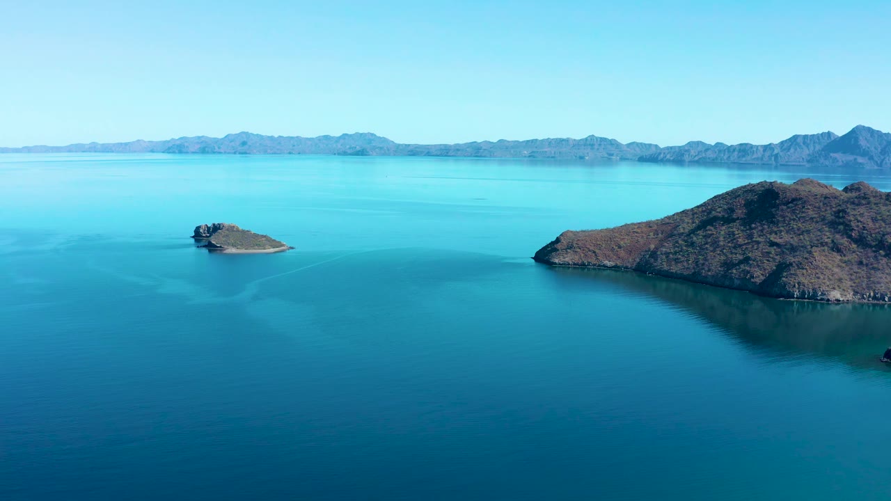 At 0:01, the small islet sits left of center while the larger scrub-covered island fills the right midground, with calm water and the Juncalito mountain horizon behind it, and no visible people.