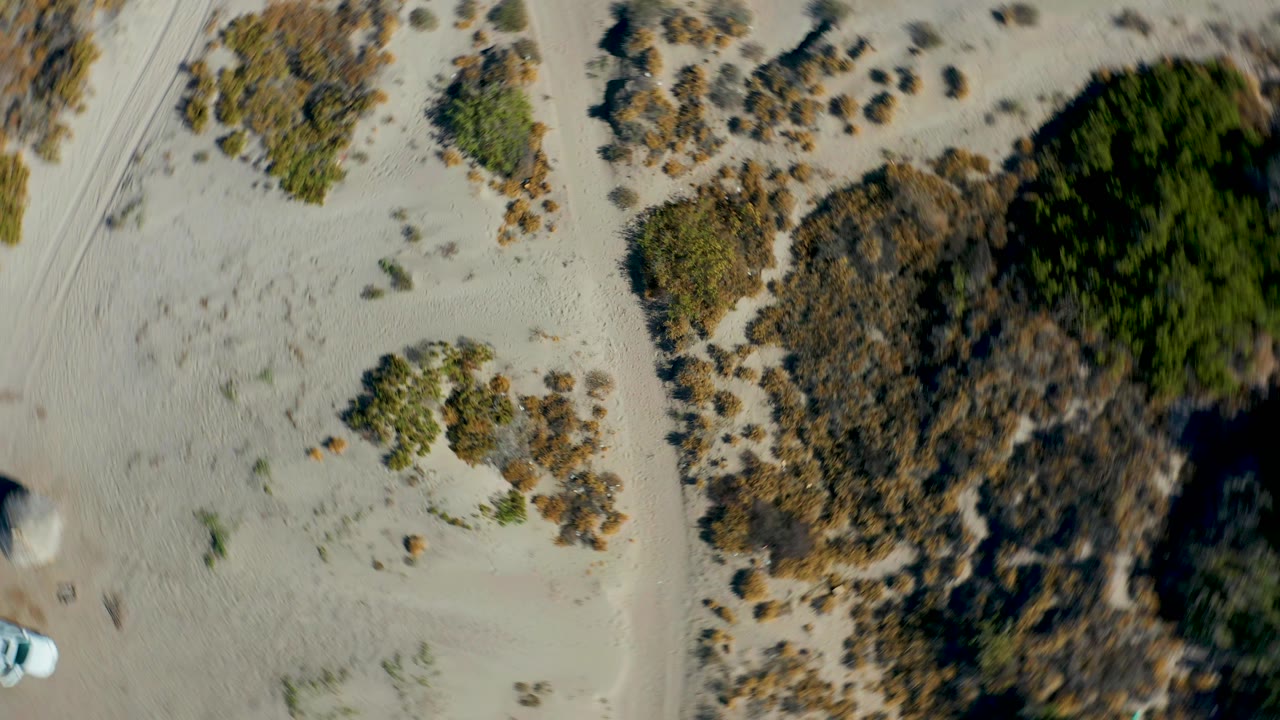 Aerial frame in Juncalito showing pale dunes, darker vegetation patches, sandy tracks, and a small white vehicle near the lower left, with no visible people.