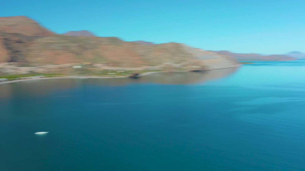 A second aerial frame over Juncalito shows calm blue ocean dominating the foreground, a faint curved beach below the mountains, a small white boat, and no visible people.