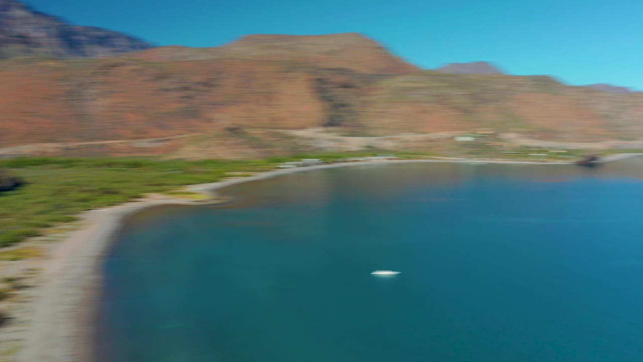A clearer aerial frame over Juncalito reveals the curved sandy beach, turquoise bay water, green vegetation at the shore, brown mountains, a small white boat, and no visible people.