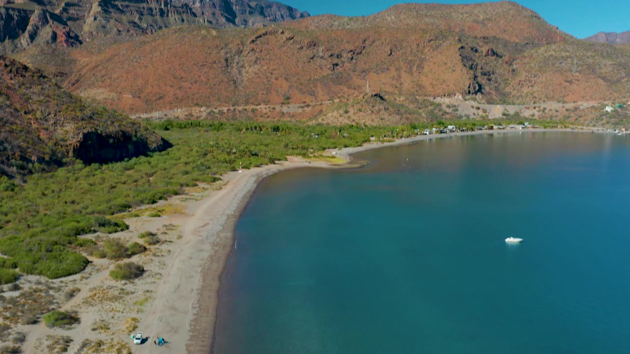 Juncalito shoreline fills the frame with a sandy beach at left, green coastal vegetation, calm blue water at right, rugged mountains behind, a small white boat, and no visible people.