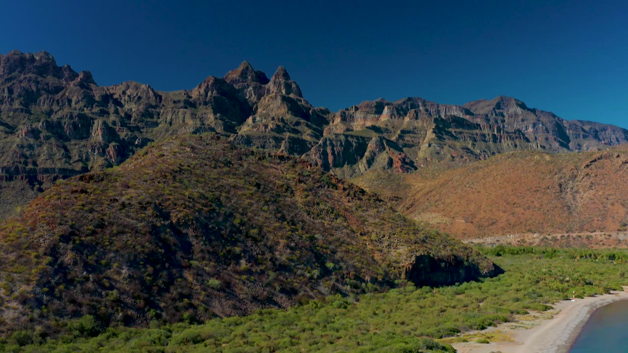Opening aerial frame over Juncalito, Baja California Sur, with jagged mountains behind a shrub-covered hill, green valley growth, a pale river corridor at right, and no visible people.