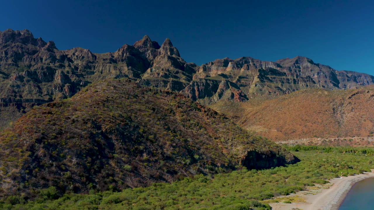 Aerial frame in Juncalito, Baja California Sur, holding on the rounded foreground hill, green valley floor, pale river channel along the right edge, rugged mountains, and no visible people.