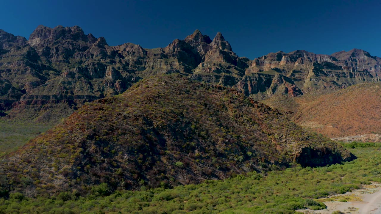 Wide Juncalito aerial frame with the foreground hill slightly lower in view, green vegetation across the valley, a pale river route to the right, jagged mountain peaks, and no visible people.