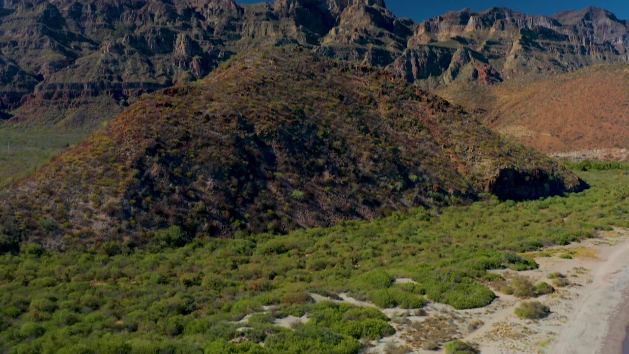 Closer aerial view of a shrub-covered hill with mountains and valley vegetation behind it.