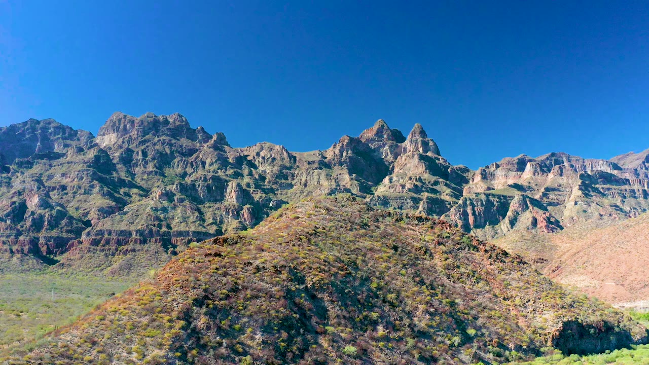 Juncalito, Baja California Sur mountain range with layered rock faces behind a dry foreground hill, clear blue sky, and no visible people at the start of the clip.