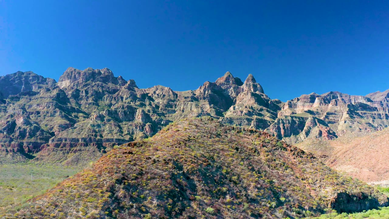 Juncalito mountain landscape one second in, showing the rounded scrub hill lower in frame, ridged brown and gray rock formations, bright sun, and no visible people.