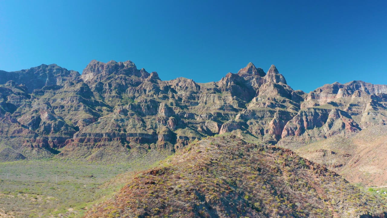 Rugged layered mountains beyond a scrub-covered desert hill under blue sky.