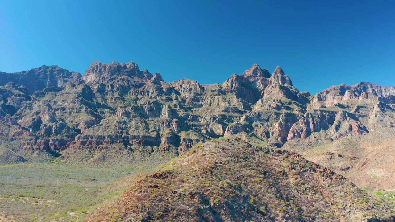 Early frame of the Juncalito mountain wall, showing layered rock faces, dry vegetation, a rounded foreground hill, and no visible people.