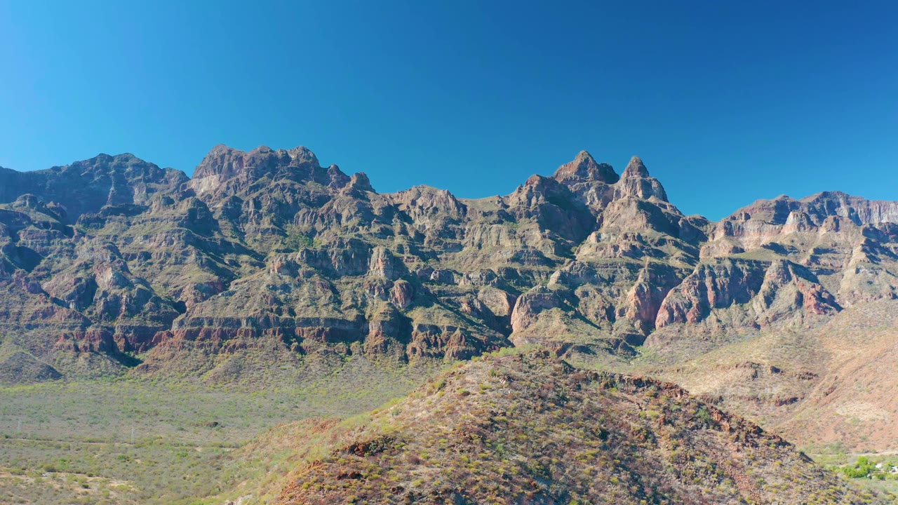 Second frame showing the same Juncalito mountain wall with tan and reddish rock bands, sparse desert vegetation across the valley, a foreground hill, and no visible people.