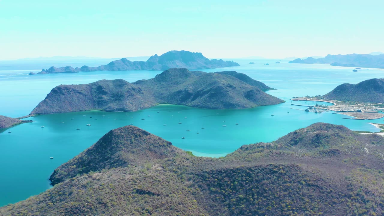 Opening aerial frame over Juncalito, Baja California Sur, showing scrub-covered island hills, clear turquoise water, scattered small boats, distant islands, and no visible people.