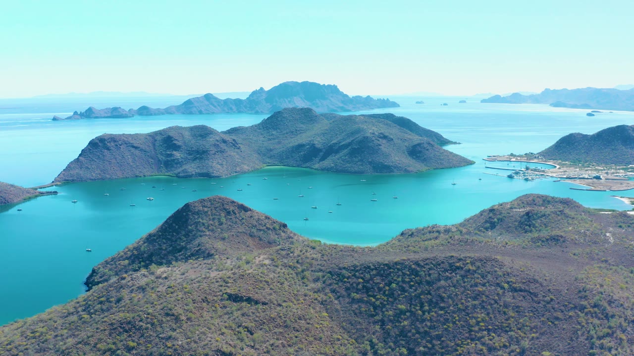 Aerial frame over Juncalito showing the rounded foreground hill lower left, calm turquoise water through the center, scattered small boats, midground islands, distant landforms, and no visible people.