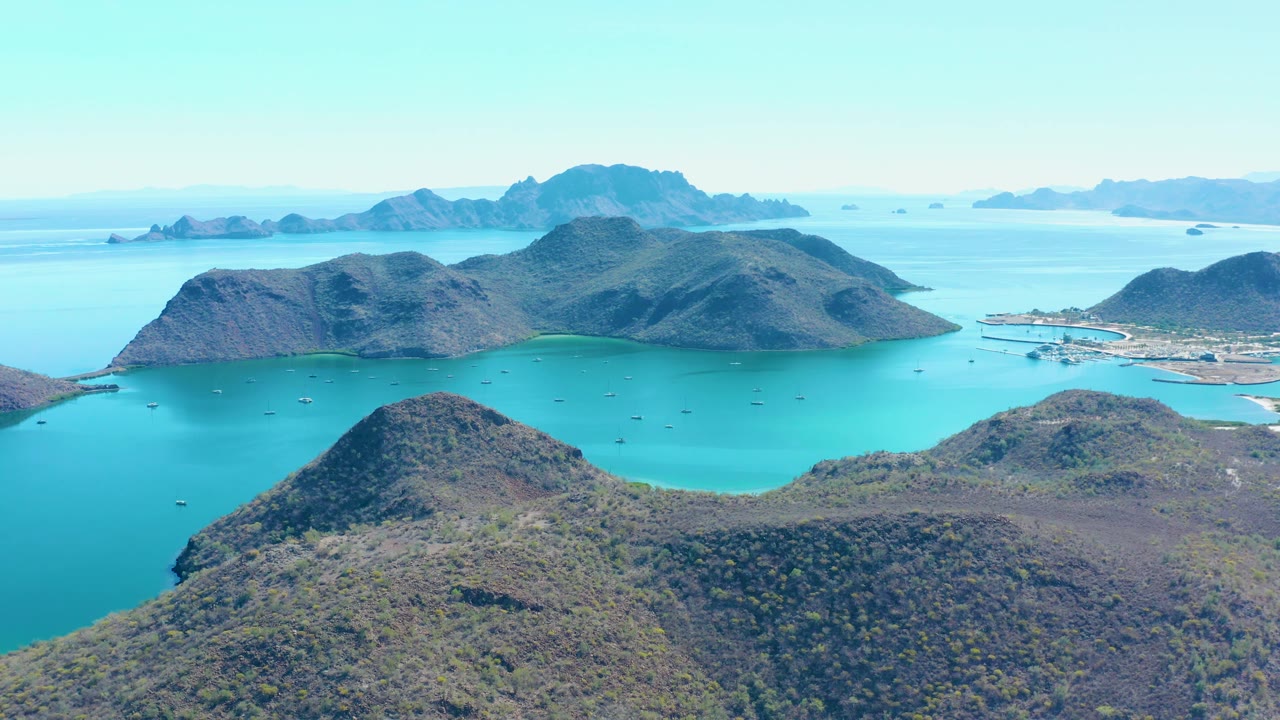 Juncalito aerial view with dry hills across the lower frame, bright turquoise bay water, several small boats, rocky islands, a small developed shoreline at right, and no visible people.