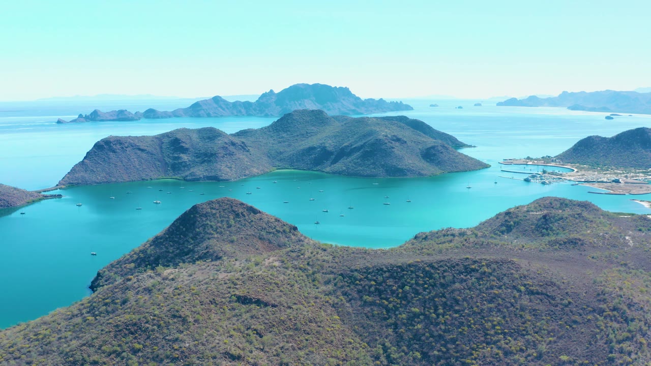 Aerial frame over Juncalito with the foreground hills filling the lower third, calm blue-green water behind them, rocky islands across the center, faint horizon islands, and no visible people.