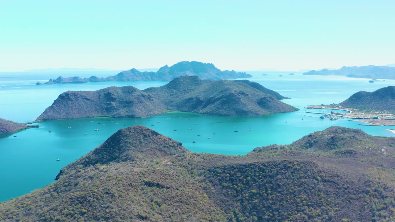 Aerial view over Juncalito, Baja California Sur, showing scrub-covered island hills, turquoise bay water, small boats, distant ridges, and no visible people.