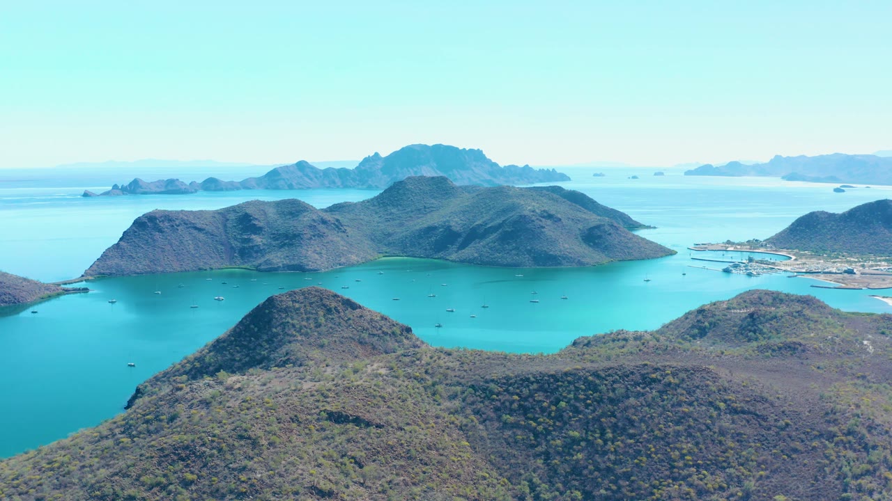 Aerial view in Juncalito, Baja California Sur, looking across a turquoise bay with small dark boats, brown-green island hills, a light hazy horizon, and no visible people.