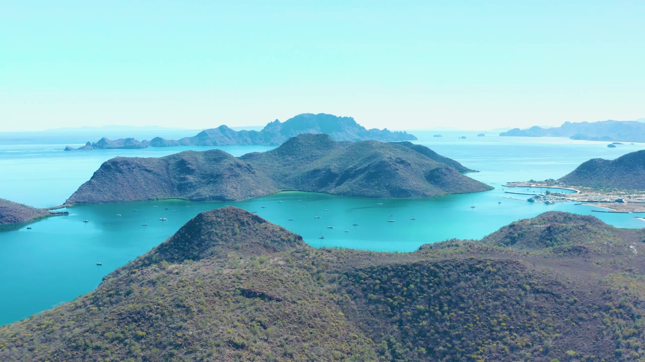 At 0:01, the Juncalito aerial view holds on scrub-covered foreground hills, bright turquoise water, island ridges across the bay, scattered small boats, and no visible people.