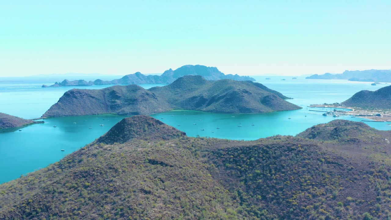 Aerial frame over Juncalito showing the foreground hillside slightly lower in view, blue-green bay water wrapping around dark rounded islands, distant mountains, and no visible people.