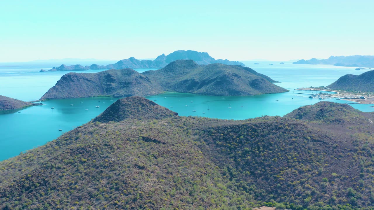 Mid-clip aerial frame over Juncalito with scrub hills in the foreground, bright turquoise water around hilly islands, pale sky, distant shoreline structures, and no visible people.