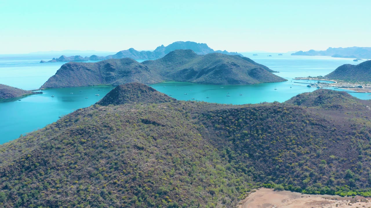 Elevated Juncalito view with a rounded foreground hill, blue-green bay water on both sides, mountainous islands beyond, and no visible people.