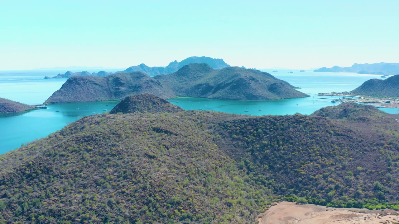 At 0:01, the aerial view over Juncalito keeps the rounded foreground hill prominent while turquoise channels and layered island silhouettes stretch across the background, with no visible people.