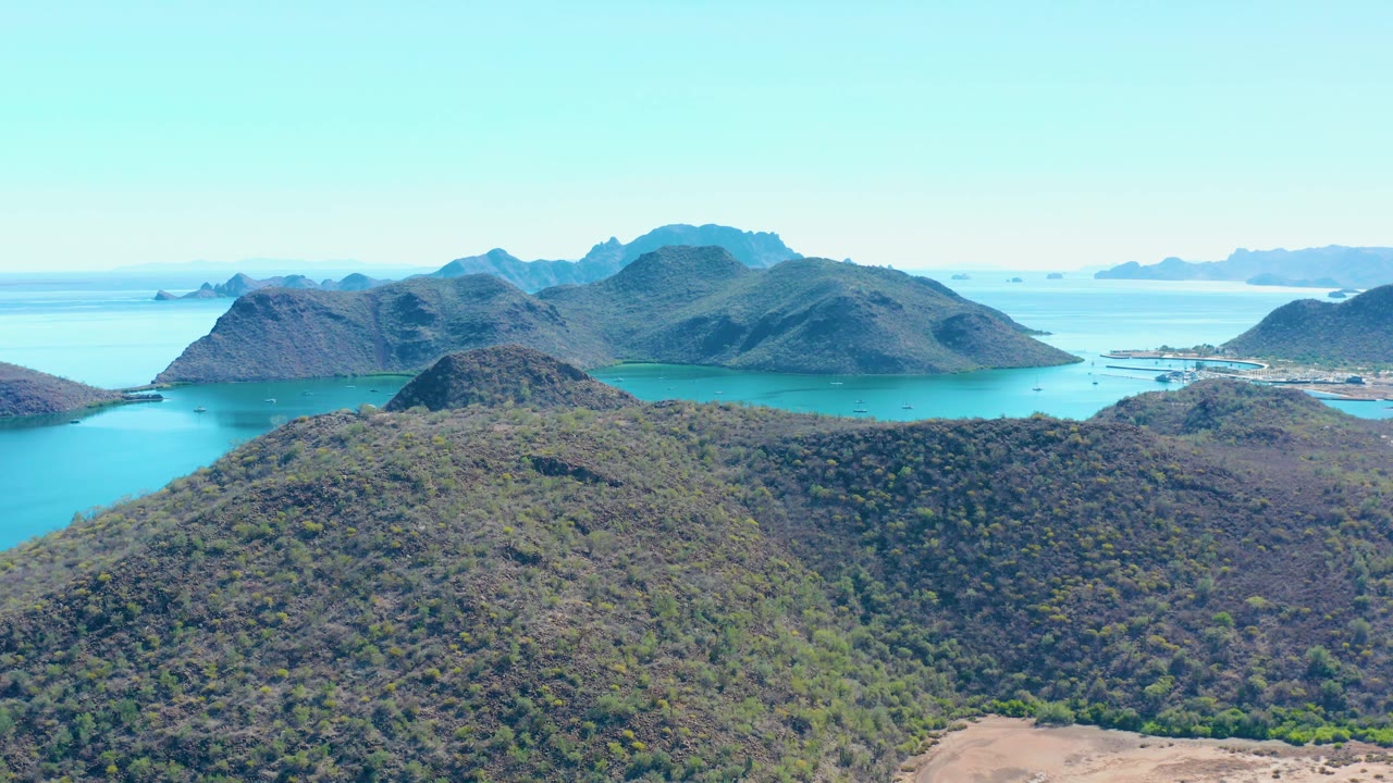 Mid-clip over Juncalito shows the foreground hillside slightly lower in frame, a turquoise inlet between hills, hazy island forms beyond, and no visible people.