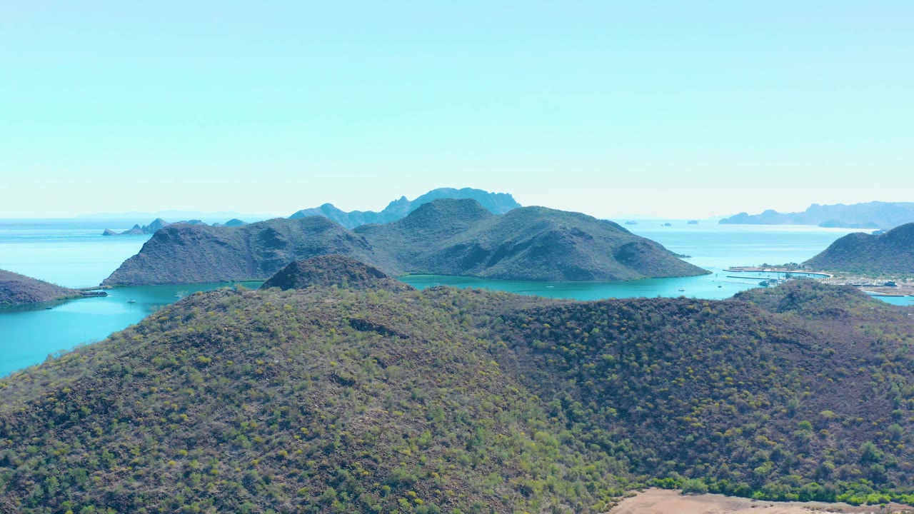 One second in, the Juncalito aerial view holds on the green foreground slope, turquoise bay channels, layered ridges, clear sky, and no visible people.