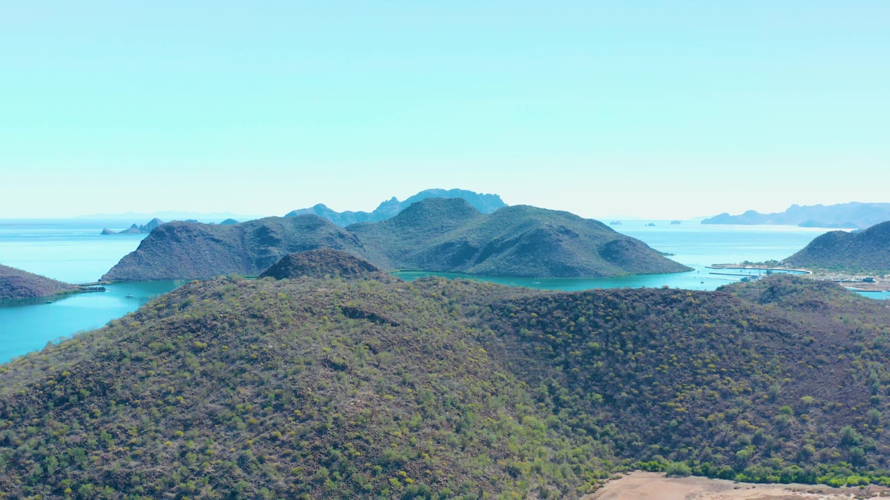 Mid-clip, the aerial view over Juncalito shows the scrubby hill rising across the lower frame, calm bay water on both sides, dark background mountains, and no visible people.