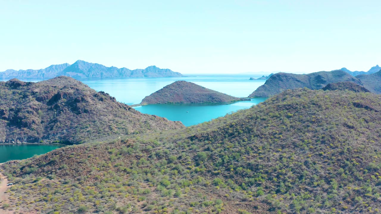 Aerial view in Juncalito showing a rounded foreground hill, a blue water channel behind it, and layered mountain ridges in the distance, with no visible people.