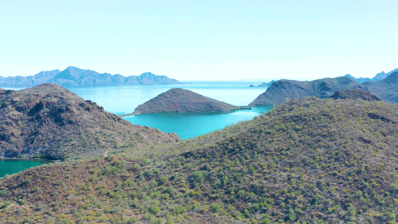 Aerial frame over Juncalito, Baja California Sur showing scrub-covered rocky hills, a turquoise inlet, distant island ridges, clear sky, and no visible people.