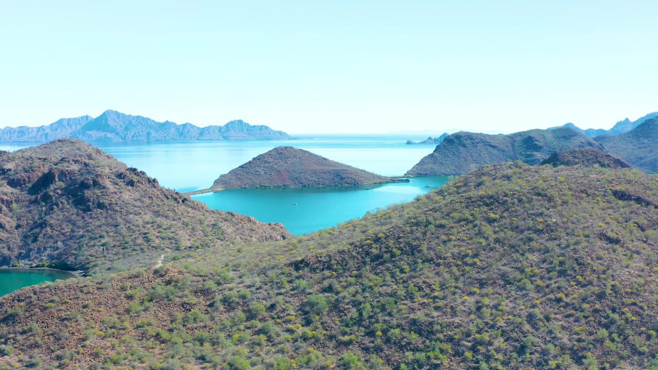 Aerial frame from Juncalito, Baja California Sur with rocky hills framing calm turquoise water, hazy blue ridges on the horizon, and no visible people.