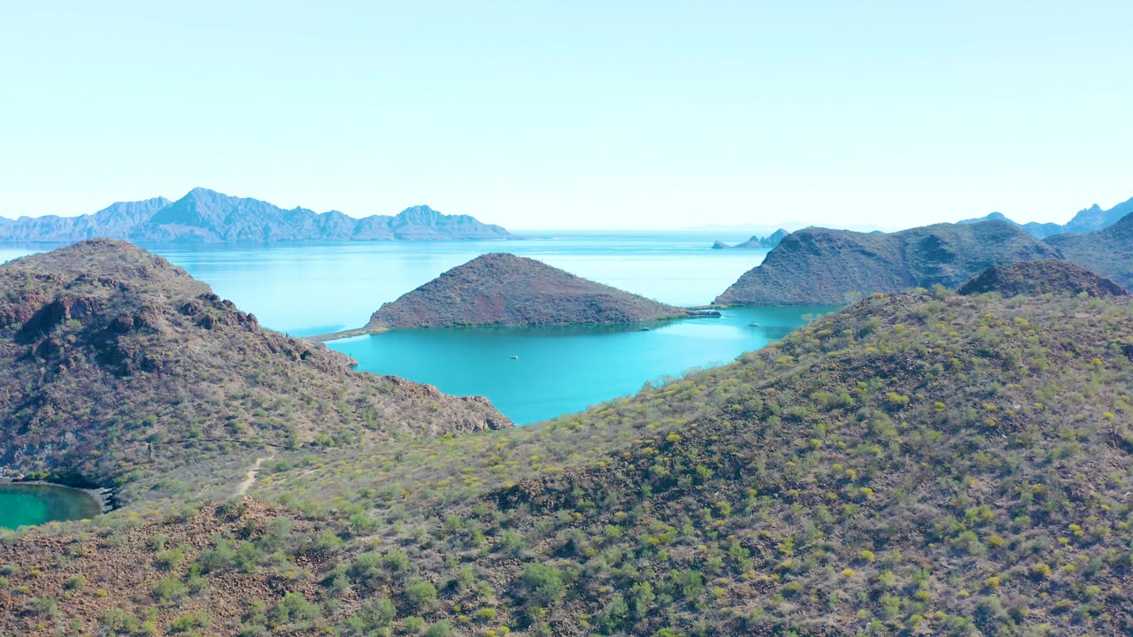 Opening aerial frame over Juncalito, Baja California Sur with dry green hills in the foreground, turquoise bay water around rocky islands, distant mountains, clear sky, and no visible people.